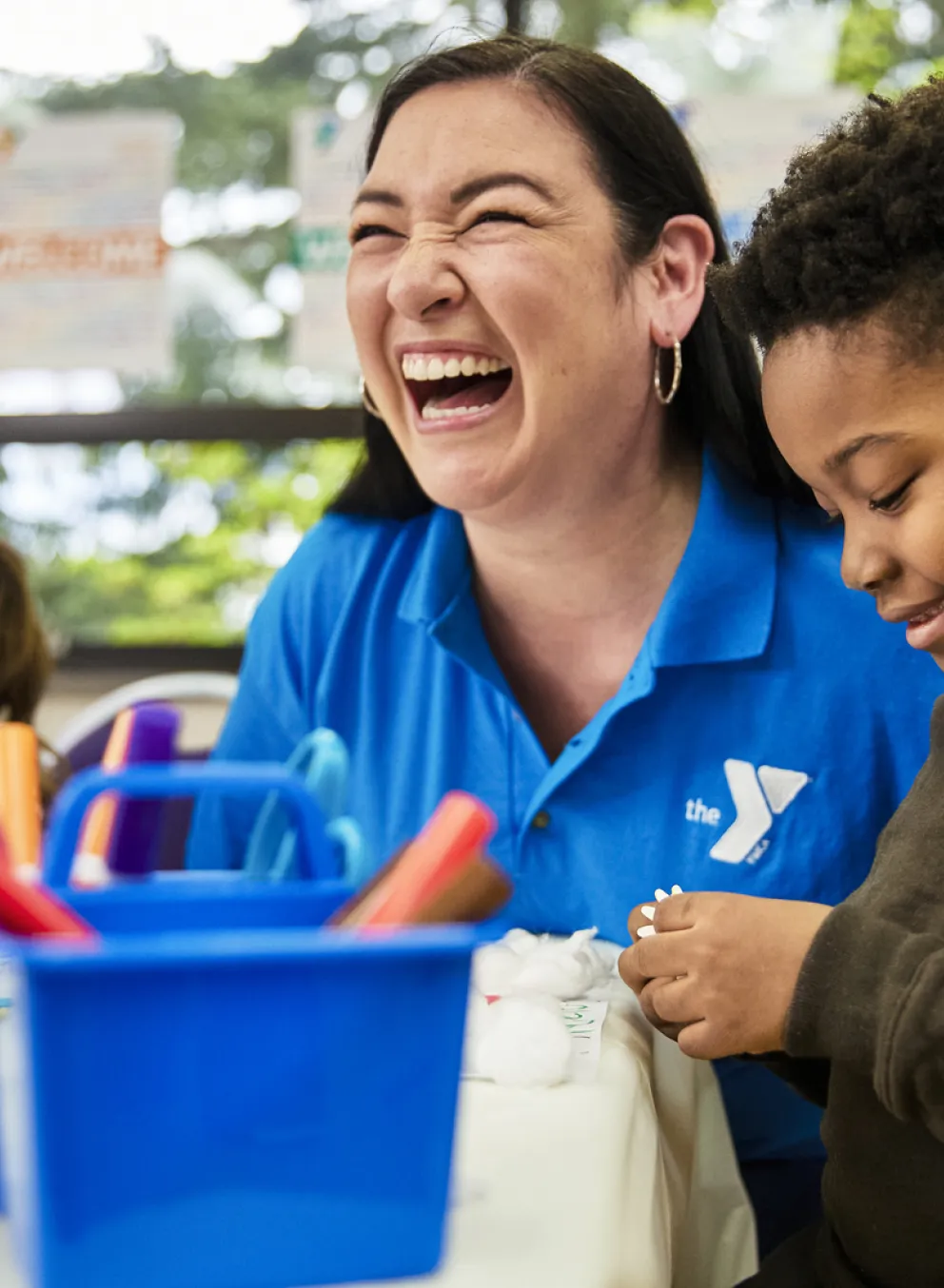 After school a Y staff member laughs and smiles while helping two young children with an art project that involves rainbows and markers; 2022 Afterschool Staff Recruitment