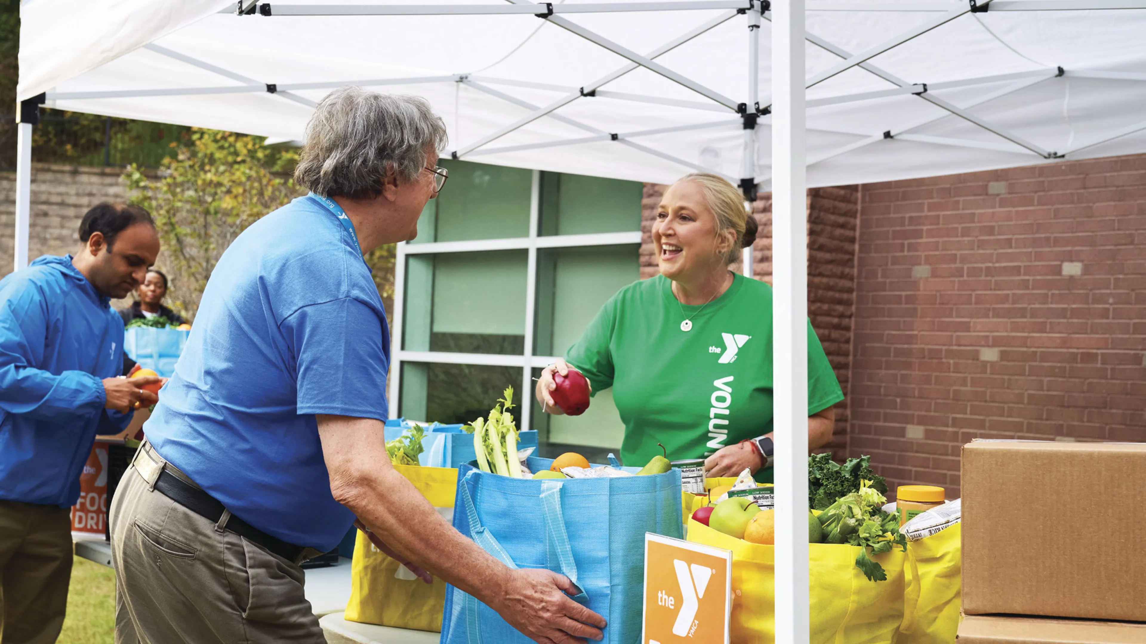 Four adults (some Y staff and some volunteers) work outdoors under a tent at a food drive. Find Your Y