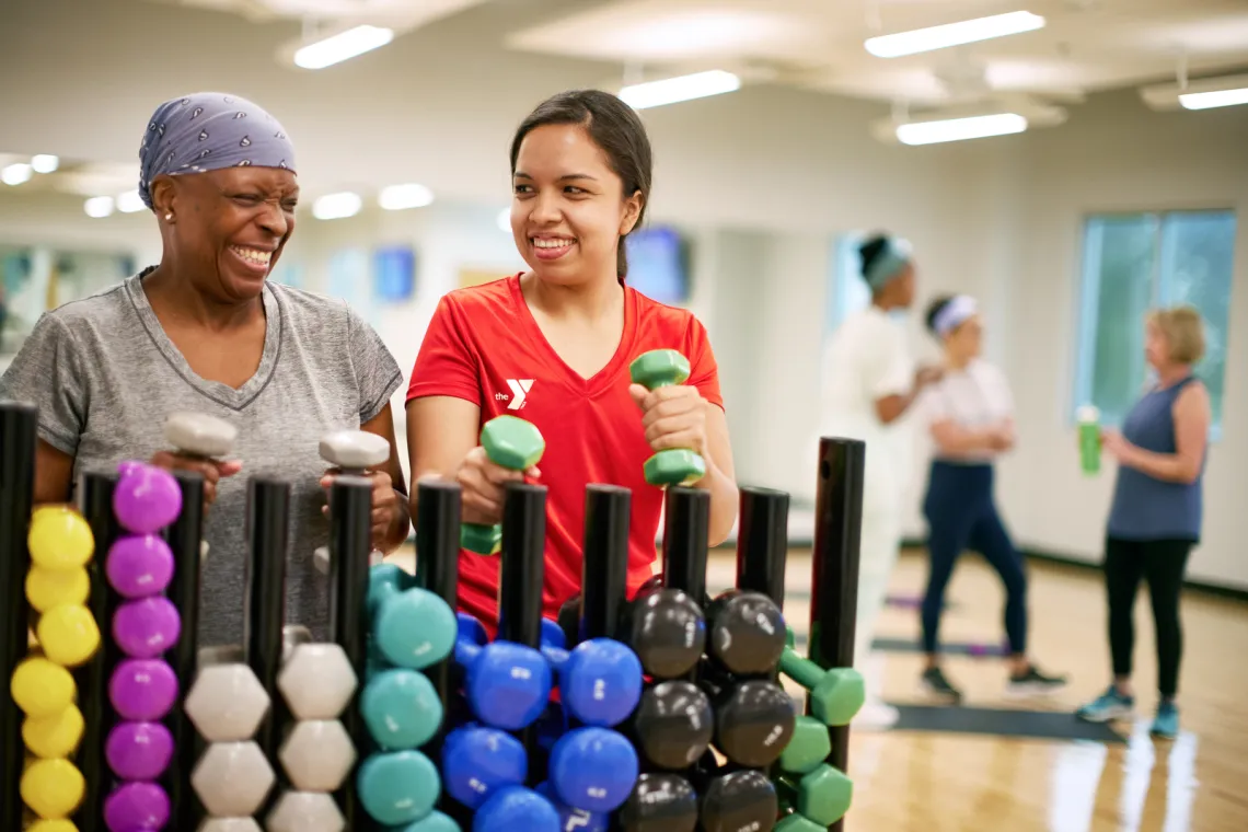 A YMCA staff member and workout class attendee smile and laugh while getting dumbbells for the workout class as the other class attendees talk in the background; Find Your Y Photography